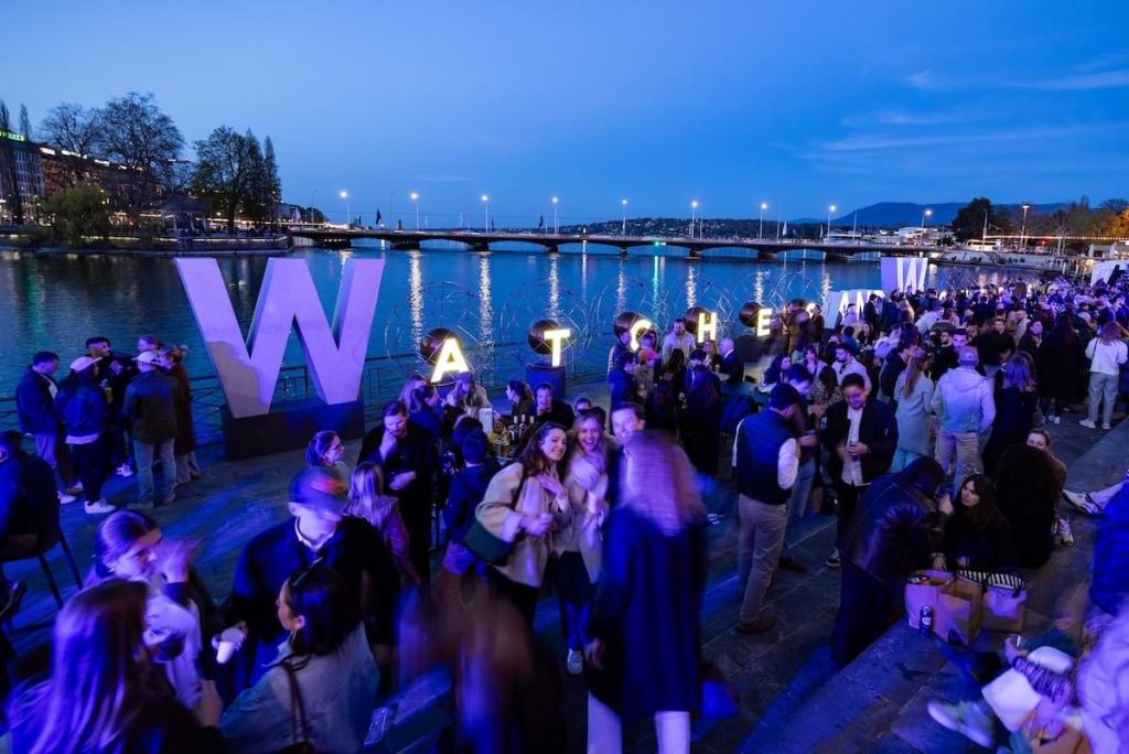 Crowd gathering at Watches and Wonders “In The City” evening event by the Geneva waterfront with illuminated signage and social atmosphere