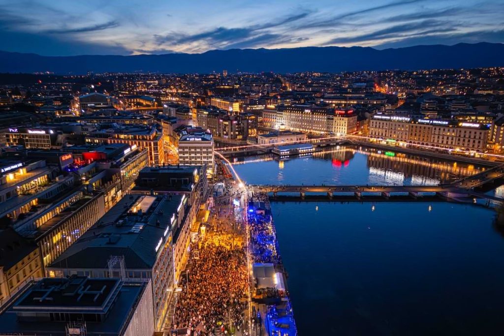 Evening aerial view of Geneva waterfront with large crowds and city lights during Watches and Wonders “In The City” events